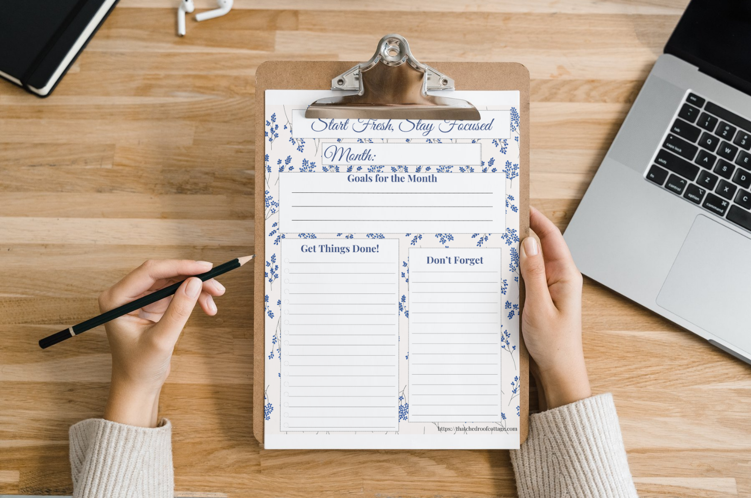 A person's hands holding a pen over a printable productivity planner with floral designs on a desk next to a laptop and papers.